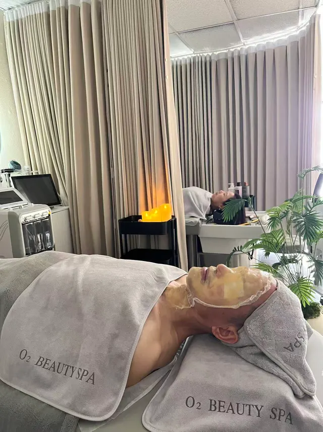A facial client resting under a gold sheet mask and gray O2 Beauty Spa towels, with a second client at a head spa basin in the background