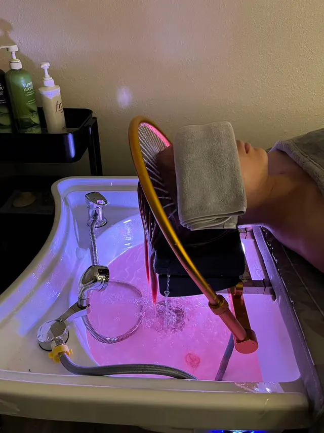Client lying back at a rose-lit head spa basin, a folded towel over the eyes, as water runs from a gold arch through the hair